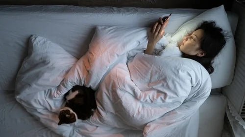 Woman lying in bed using phone with cat beside her showcasing best relaxation products 2023 for comfort